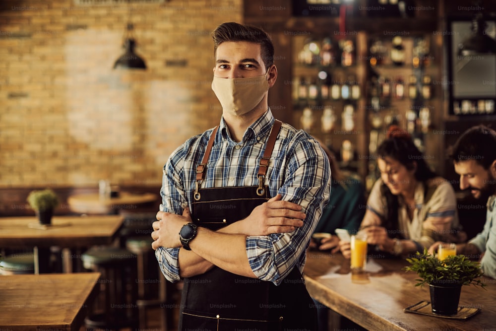 Porträt eines Kellners mit Schutzmaske, der mit verschränkten Armen in einer Kneipe steht und in die Kamera schaut.