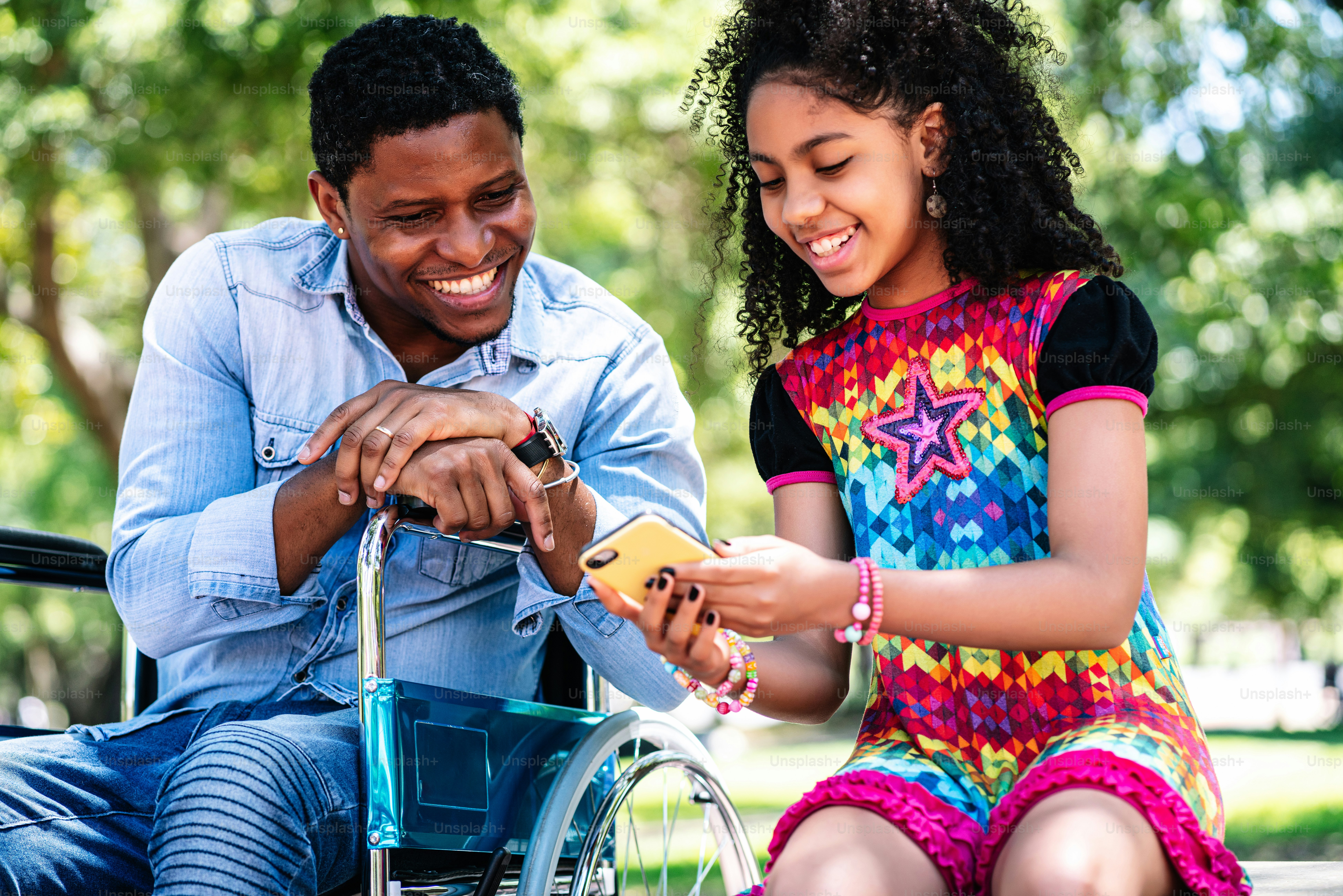 Un hombre afroamericano en silla de ruedas usando un teléfono móvil con su hija mientras disfrutan de un día juntos en el parque.