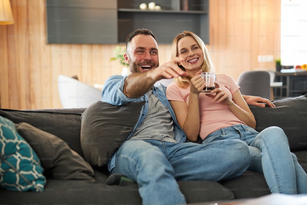 Charming woman holding cup of coffee and smiling while husband pointing at something