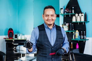 Retrato del barbero del hombre latino sosteniendo los equipos en la mano, mirando a la cámara en una pequeña empresa de la barbería en la ciudad de México