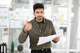 Handsome indian man in casual wear showing thumb up on camera. Young creative office worker holding papers and gesturing indoors.