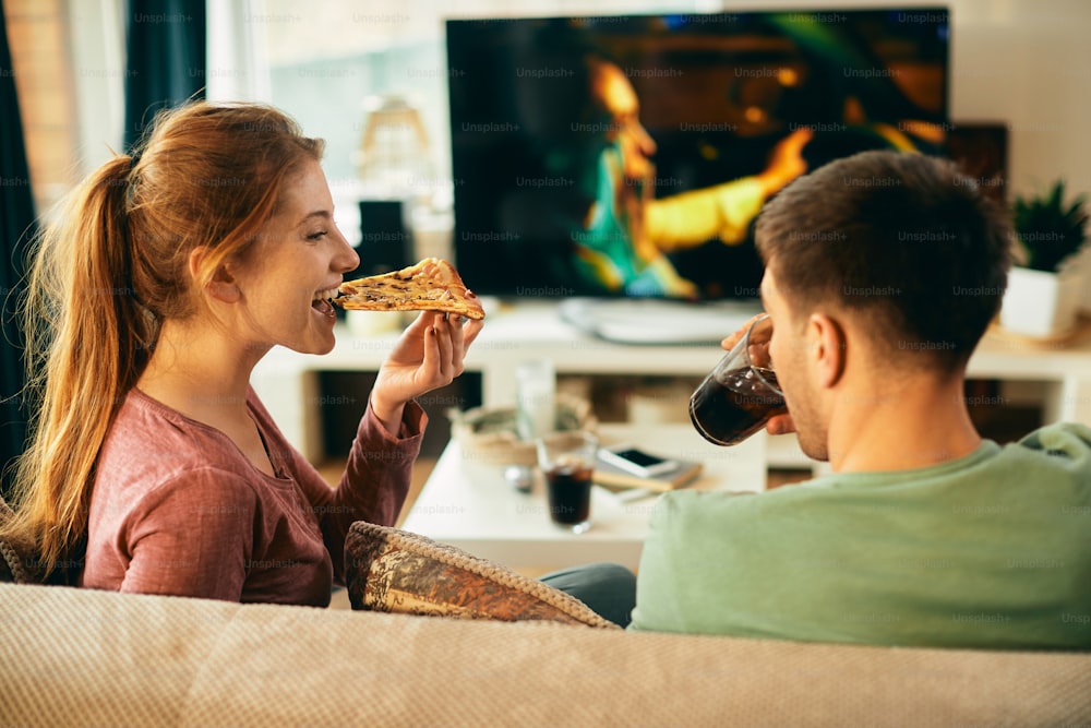 家でテレビを見ながらくつろぐ若いカップル。焦点はピザを食べる女性です。