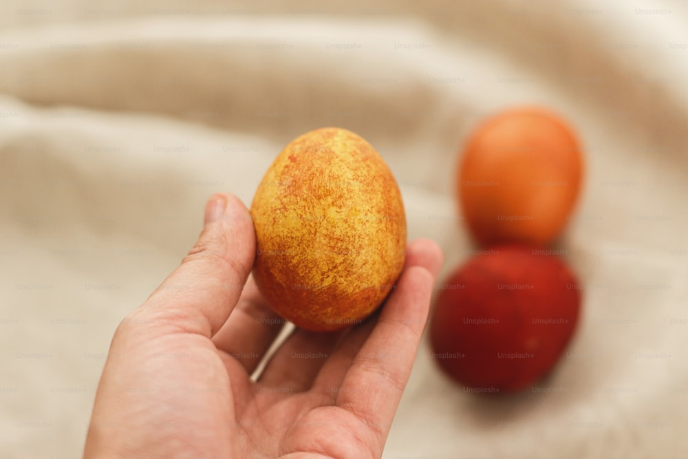 Hand holding modern easter egg on rustic linen cloth background. Happy Easter! Natural hand made dyed eggs in red and yellow color on grey textile. Aesthetic