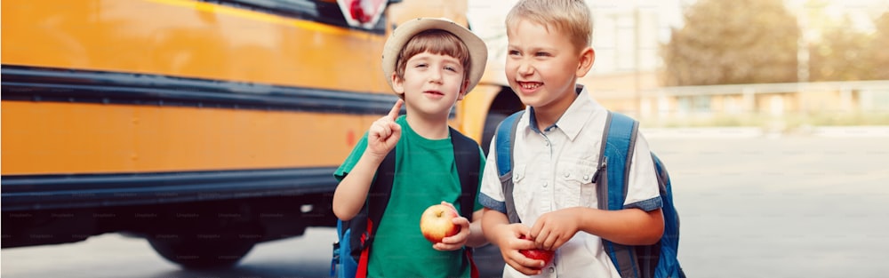 Two funny happy Caucasian boys students kids with apples standing by yellow bus on 1 September day. Education back to school. Children ready to learn and study. Web banner header for website.