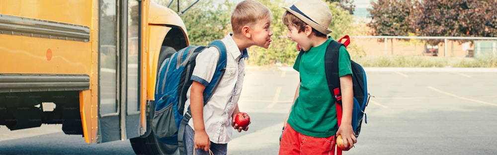Dois engraçados meninos caucasianos felizes mostrando línguas um ao outro perto de ônibus amarelo no dia 1 de setembro. Educação de volta às aulas. As crianças aprendem e estudam. Cabeçalho do banner da Web para o site.