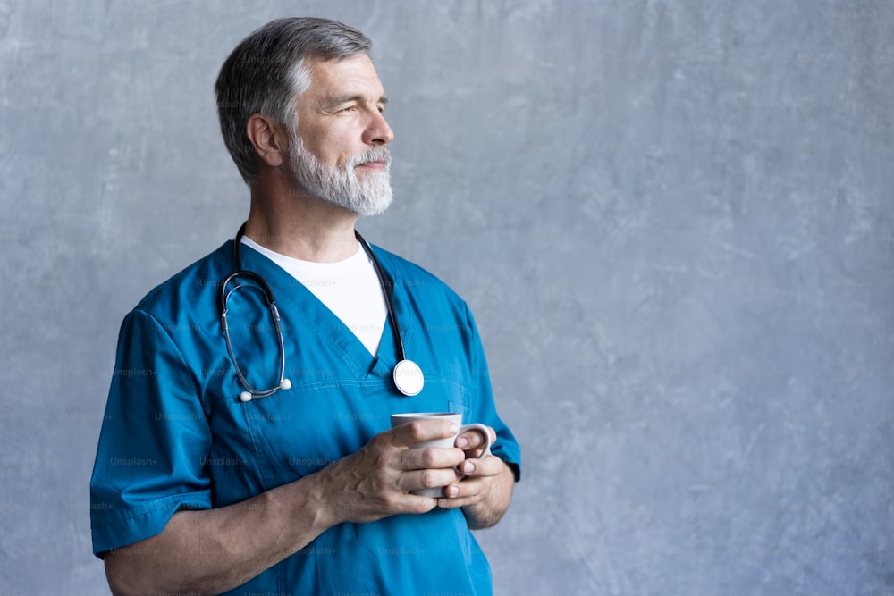 Retrato del cirujano maduro profesional que sostiene la taza, mirando a la cámara mientras está de pie contra el fondo gris