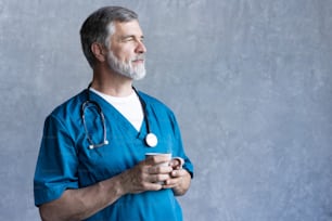 Portrait of professional mature surgeon holding cup, looking at camera while standing against the grey background