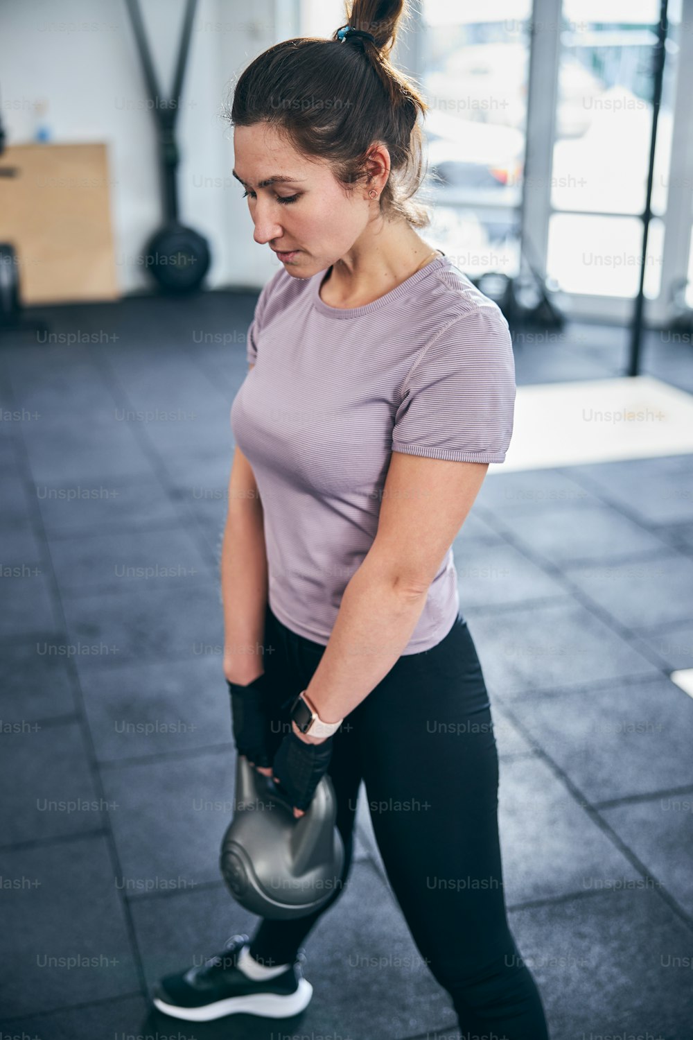 Bodybuilder femminile caucasico concentrato calmo in guanti da allenamento senza dita che tiene un kettlebell con entrambe le mani