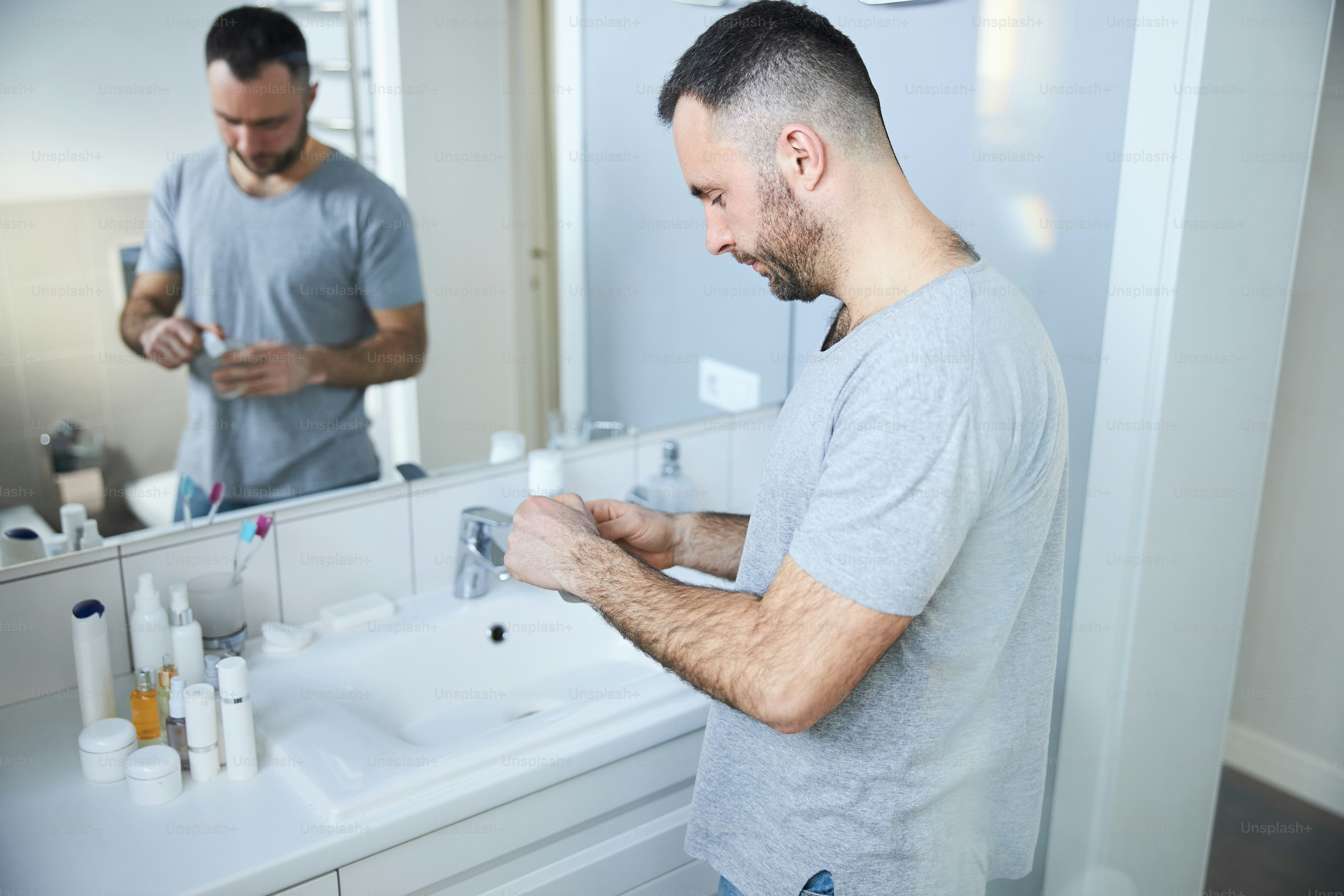 Bearded male person holding bottle of cologne while standing by the sink in front of the mirror