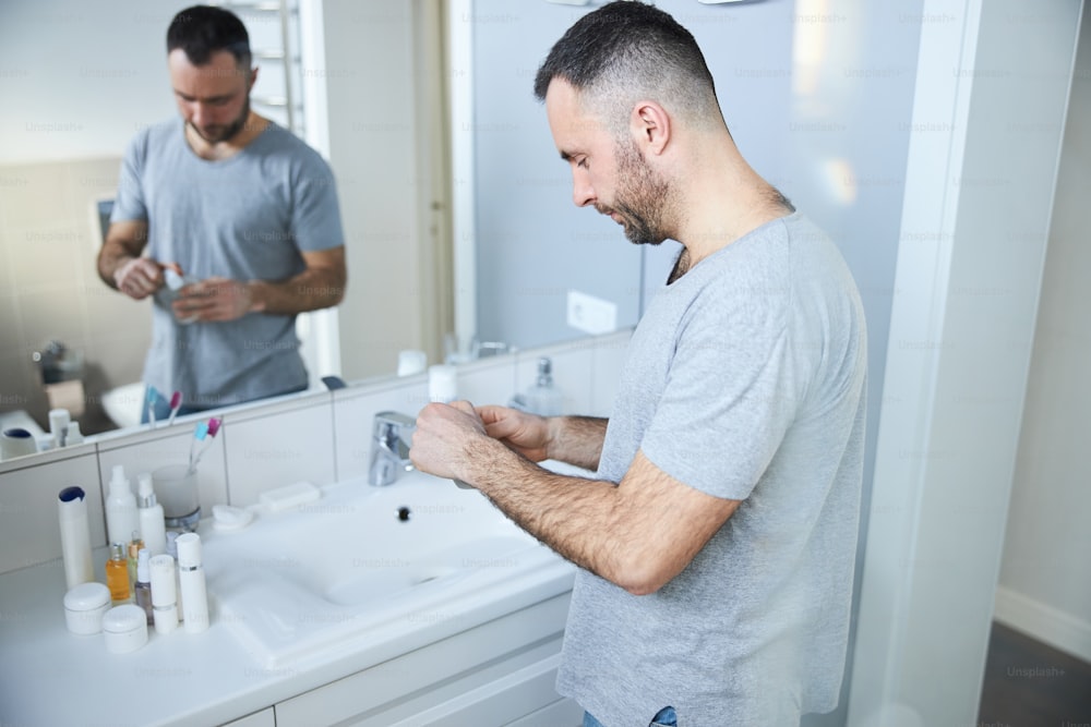 Persona maschio barbuta che tiene una bottiglia di acqua di colonia mentre si trova vicino al lavandino davanti allo specchio