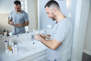 Bearded male person holding bottle of cologne while standing by the sink in front of the mirror