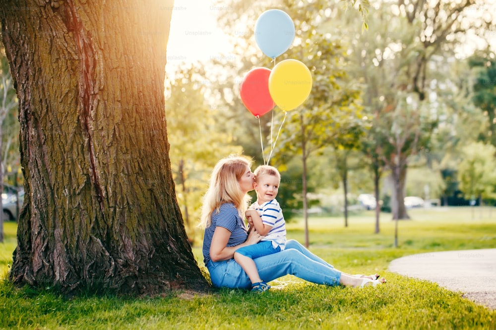 Young Caucasian mother and boy toddler son sitting together with balloons in park. Family mom kissing child outdoor on summer day. Happy authentic family childhood lifestyle.