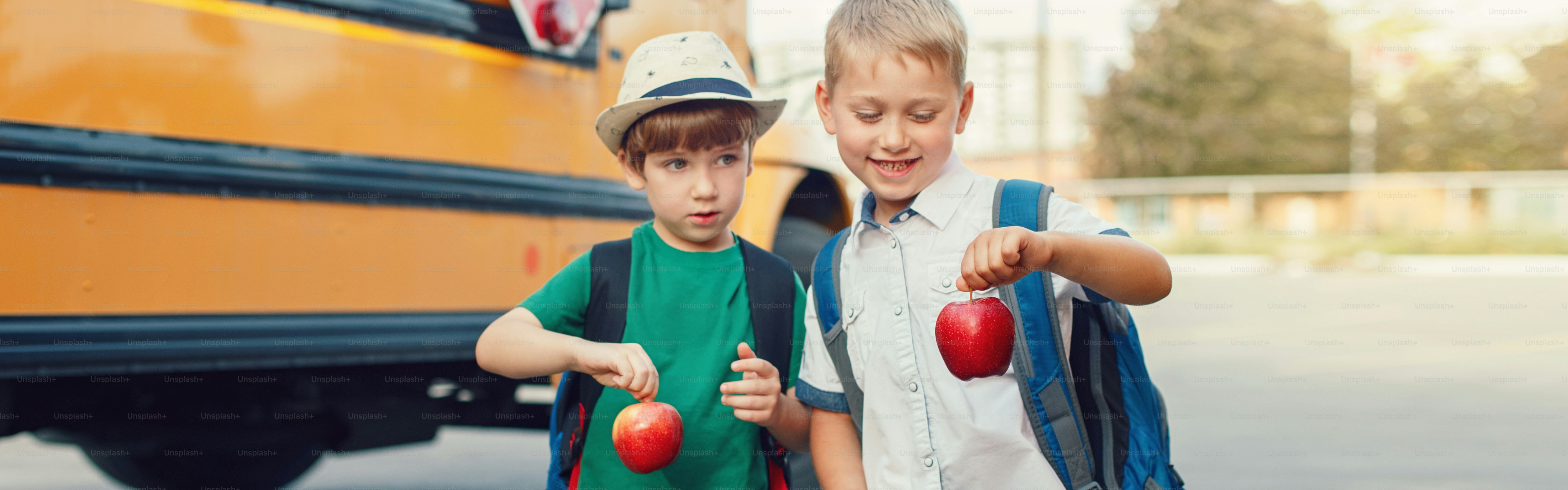 Dos divertidos niños caucásicos felices estudiantes niños con manzanas de pie junto al autobús amarillo el día 1 de septiembre. Regreso a la escuela de la educación. Niños listos para aprender y estudiar. Encabezado de banner web para sitio web.