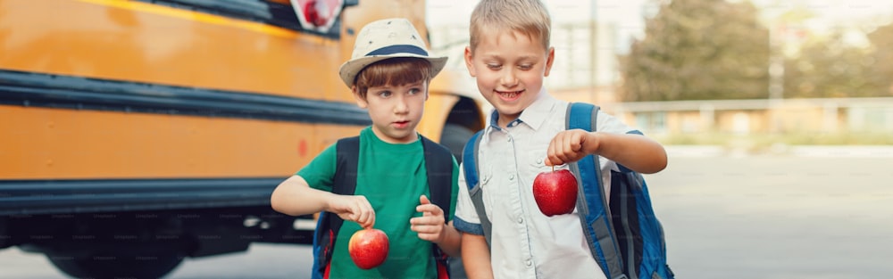 Dois engraçados meninos caucasianos felizes estudantes crianças com maçãs de pé ao lado de ônibus amarelo no dia 1 de setembro. Educação de volta às aulas. Crianças prontas para aprender e estudar. Cabeçalho do banner da Web para o site.