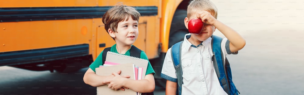 Two funny happy Caucasian boys students kids with apples standing by yellow bus on 1 September day. Education back to school. Children ready to learn and study. Web banner header for website.