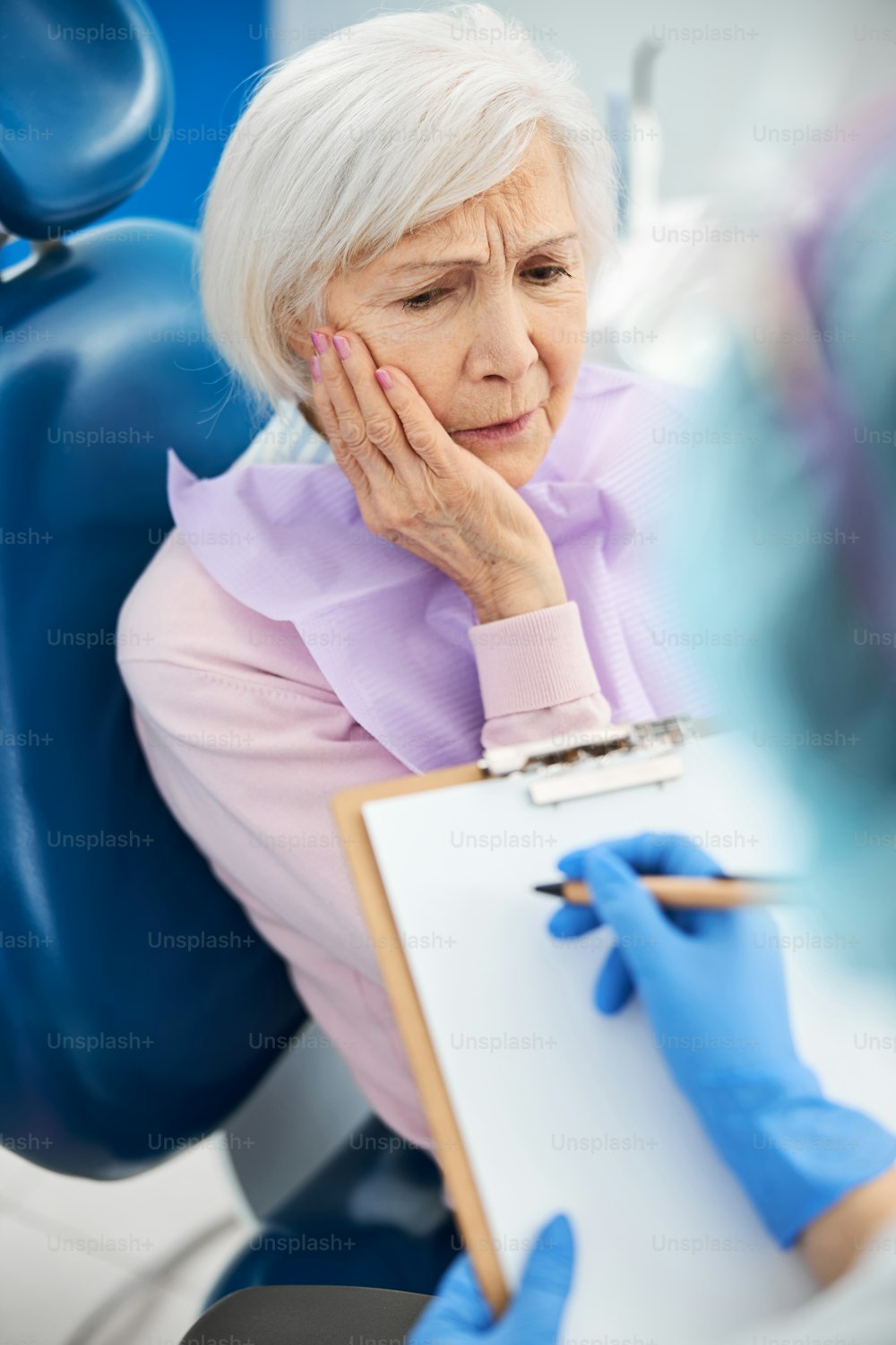 Señora anciana molesta presionando su palma contra la mejilla derecha mientras le cuenta sobre sus problemas dentales a una persona que toma notas