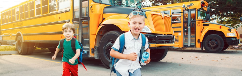 Two funny happy Caucasian boys students kids running near yellow bus on 1 September day. Education back to school. Children ready to learn, study. Web banner header for website.