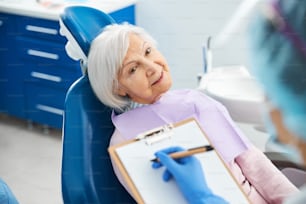 Alternde Dame in einem Zahnarztsessel beobachtet, wie der Arzt Notizen in einem Notizbuch mit Öko-Stift macht