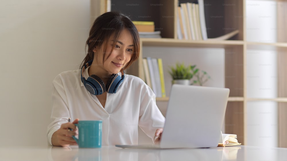Ritratto di freelance femminile con cuffia che tiene la tazza di caffè mentre lavora con il computer portatile nella stanza dell'ufficio di casa