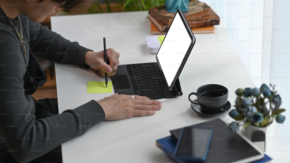 Side view of young man freelancer using computer tablet and making note on sticky note at home office.