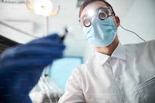 Médecin masculin dans des loupes binoculaires avec une lumière vive portant un masque tendant vers le bas avec une perceuse dentaire