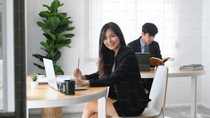 Fröhliche Büroangestellte, die in die Kamera lächelt und mit ihrer Kollegin im Büro sitzt.
