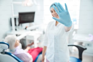 Mulher que usa luvas e chapéu descartável colocando a mão na frente de si mesma enquanto está em pé no consultório do dentista