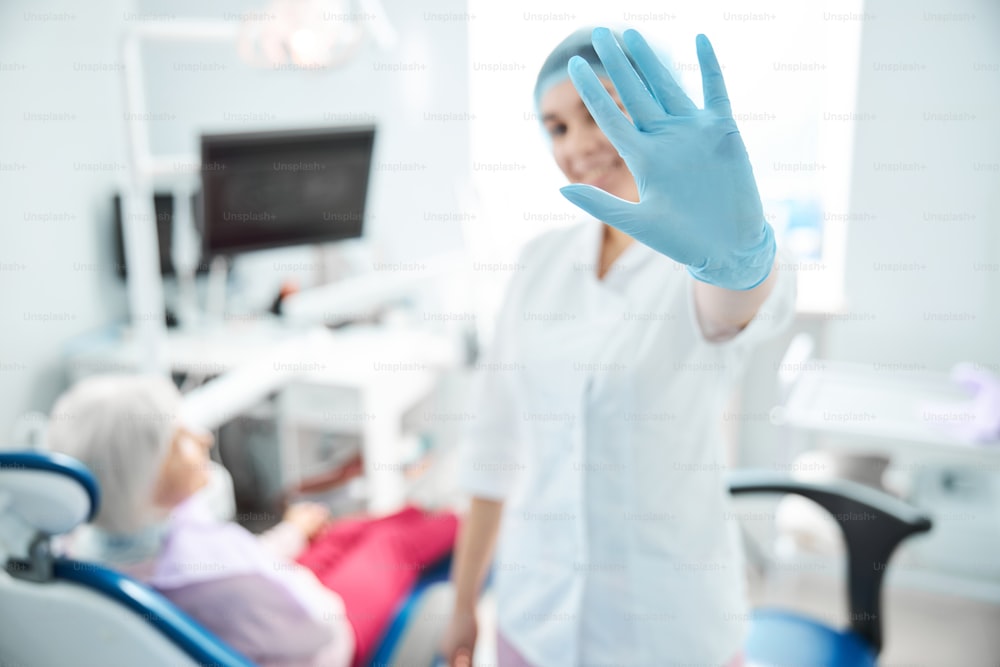 Frau mit Handschuhen und Einwegmütze hält sich die Hand vor, während sie in der Zahnarztpraxis steht