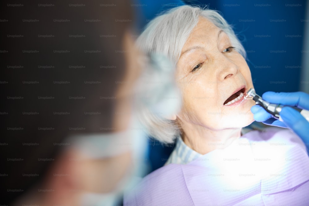Aging person with grey hair lying with opened mouth while dental drill approaching her tooth