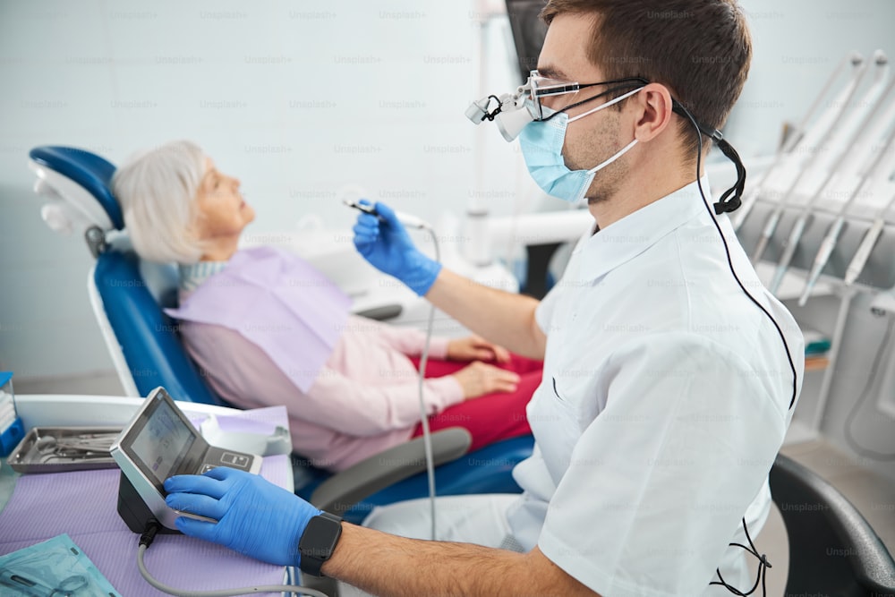 Operaio della clinica dentale in guanti medici blu e maschera che aziona un'unità di controllo per il manipolo vicino a un paziente