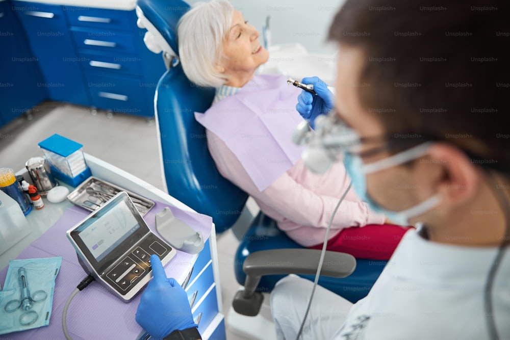 Zahnspezialist konfiguriert die Einstellungen seines Handstücks auf einem elektronischen Gerät mit älterer Dame neben ihm