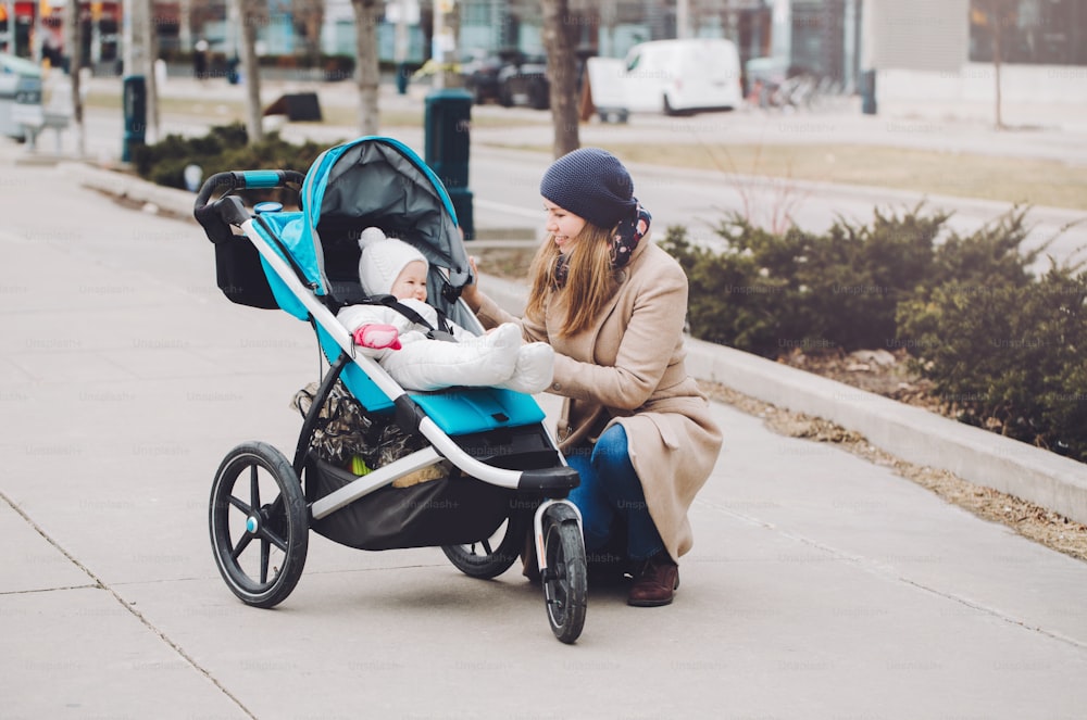 Happy young Caucasian mother walking with baby daughter outdoor in city. Family two people strolling together outside on town streets. Urban life with kids children.
