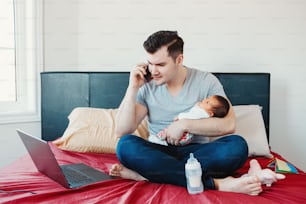 Busy Caucasian father with newborn baby working online from home. Man parent holding child talking over phone. Single dad family. Coronavirus quarantine social distance and self isolation.