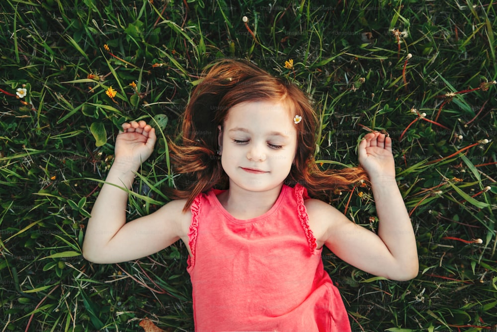 초원의 풀밭에서 쉬고 있는 꿈을 꾸는 귀여운 백인 소녀. 아이는 땅에 누워 자고 있습니다. 야외 활동 재미 여름 어린이 활동.  행복한 어린 시절 생활 방식. 위에서 본 모습.
