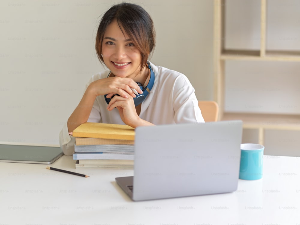 Portrait of female student smiling to camera while sitting at study table wit laptop and stack of books in living room