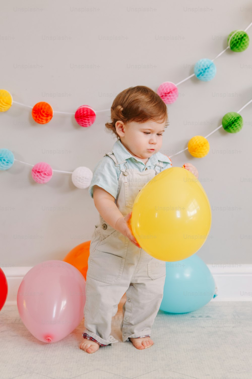 Funny Caucasian baby boy celebrating his first birthday. Excited child kid toddler playing with colorful balloons. Celebration of event or party indoors at home. Happy birthday lifestyle concept.