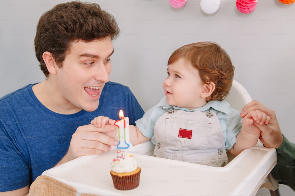 Smiling Caucasian father with baby boy celebrating his first birthday at home. Proud parent dad together with child kid toddler. Cupcake food with one lit candle. Happy birthday lifestyle concept.