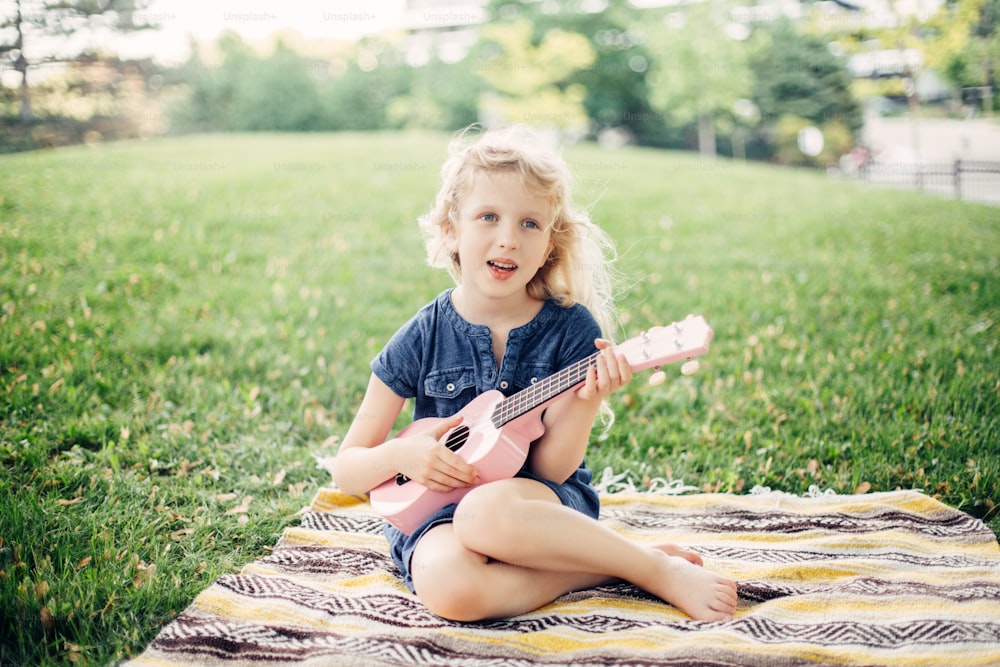 かわいい愛らしいブロンドの女の子が屋外でピンクのギターのおもちゃを演奏しています。公園で音楽を奏でたり、歌を歌ったりする子供。子供のための趣味活動。優しく、思い出に残る、本物の率直な子供時代の瞬間。