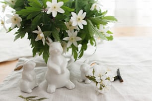 Simpatici coniglietti bianchi e fiori primaverili su tovagliolo di stoffa di lino su tavolo rustico in luce soffusa. Buona Pasqua! Concetto di caccia pasquale. Figurine di coniglio bianco e anemoni in fiore fiori natura morta rurale
