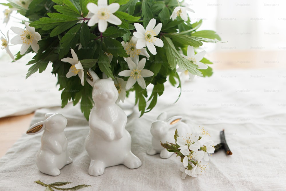 Cute white bunnies and spring flowers on linen cloth napkin on rustic table in soft light. Happy Easter ! Easter hunt concept. White rabbit figurines and blooming anemones flowers rural still life