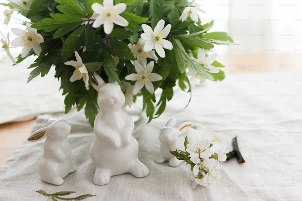 Niedliche weiße Hasen und Frühlingsblumen auf Leinentuchserviette auf rustikalem Tisch in sanftem Licht. Frohe Ostern! Osterjagd-Konzept. Weiße Kaninchenfiguren und blühende Anemonen Blumen bäuerliches Stillleben