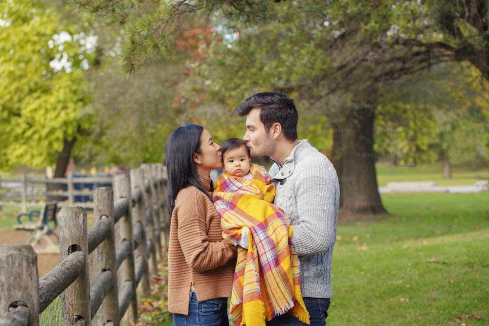 Asian Chinese mother and Caucasian father dad holding kissing baby girl wrapped in blanket. Family in autumn fall park outdoor with tree leaves nature. Seasonal holiday Thanksgiving. Family Day.