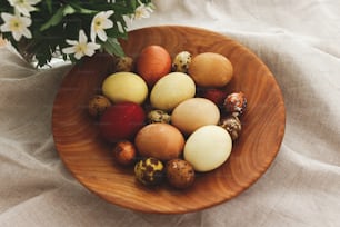 Modern easter eggs in wooden bowl on rustic linen cloth with spring flowers. Happy Easter! Natural dyed eggs in yellow and red colors on rural table with blooming flowers anemone. Aesthetic
