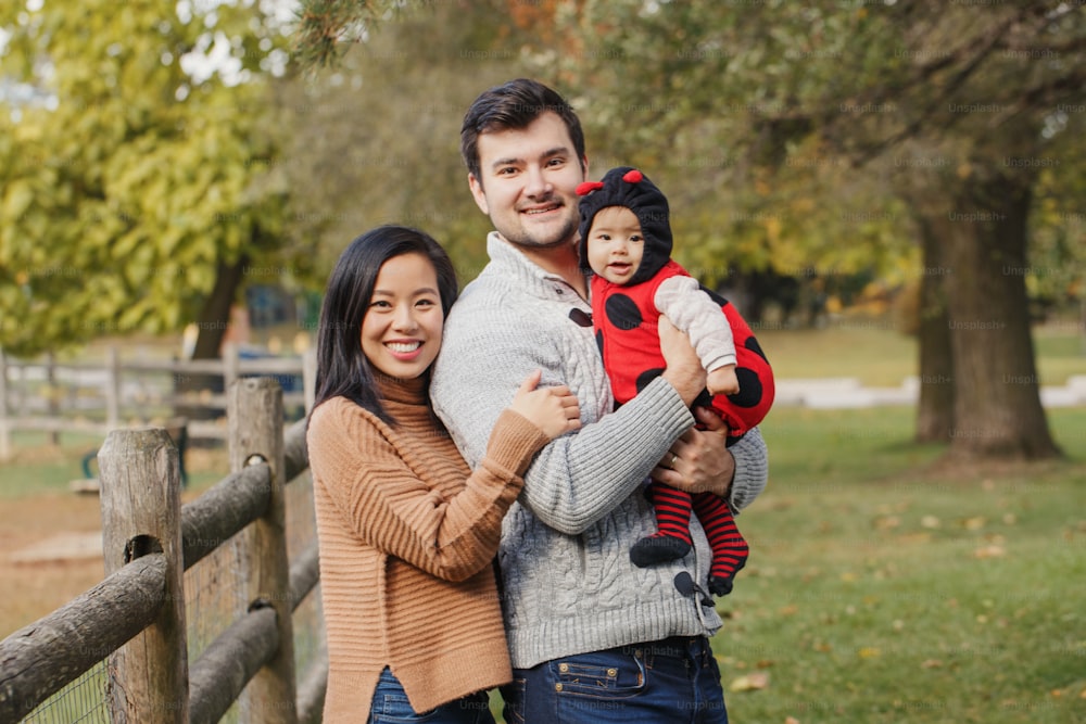 Happy smiling Asian Chinese mother and Caucasian father dad with baby girl in ladybug costume. Family in autumn fall park outdoor. Halloween or Thanksgiving holiday concept.