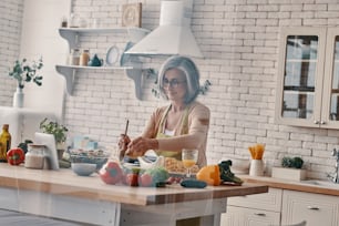 家で時間を過ごしながら健康的な夕食を作るエプロンで忙しい年配の女性