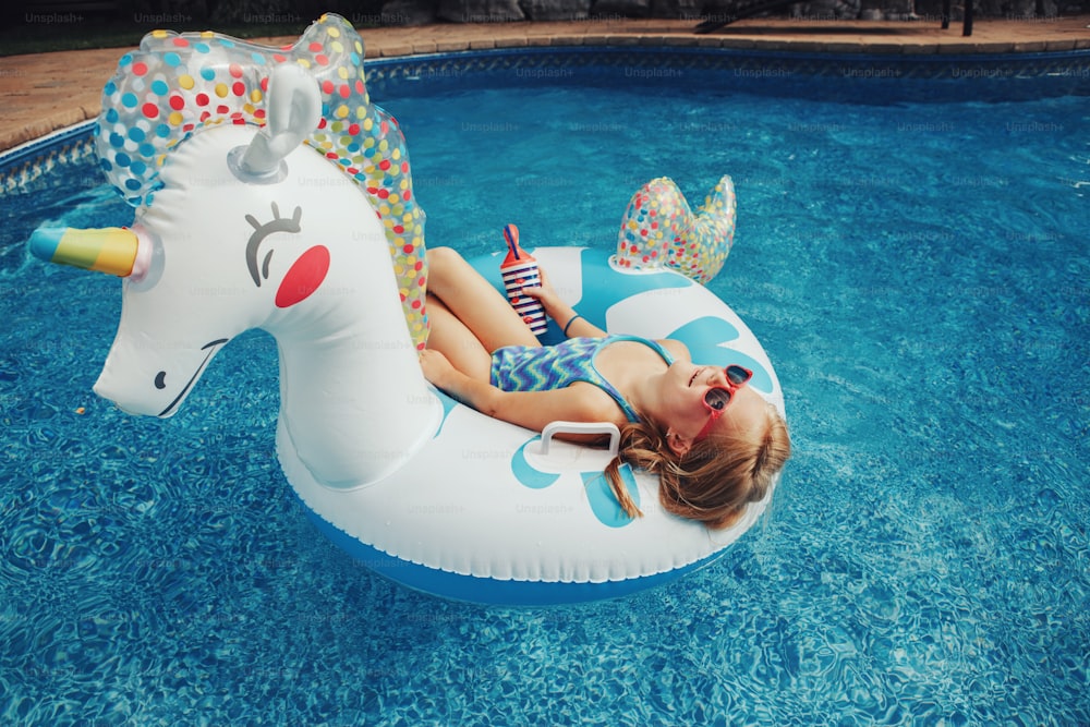 Cute adorable girl in sunglasses with drink lying on inflatable ring unicorn. Kid child enjoying having fun relaxing resting in swimming pool on floatie. Summer outdoor water activity for kids.
