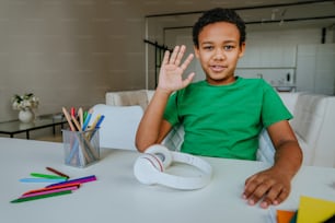 집에서 온라인 수업 중에 손을 흔들고 인사하는 카메라를 보는 소년의 초상화.