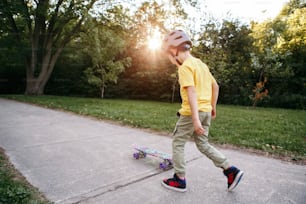 Ragazzo in casco grigio che cavalca lo skateboard sulla strada nel parco il giorno d'estate. Sport stagionale per bambini all'aperto. Stile di vita infantile sano. Ragazzo che impara ad andare in skateboard. Vista da dietro.