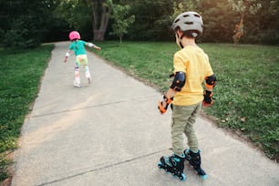 Friends boy and girl in helmets riding on roller skates in park on summer day. Sister encourage stimulate brother to ride on roller skates. Siblings rivalry. Seasonal outdoor children activity sport.