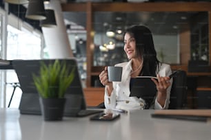 Thoughtfully businesswoman in earphone relaxing in office holding cup of coffee looking away.